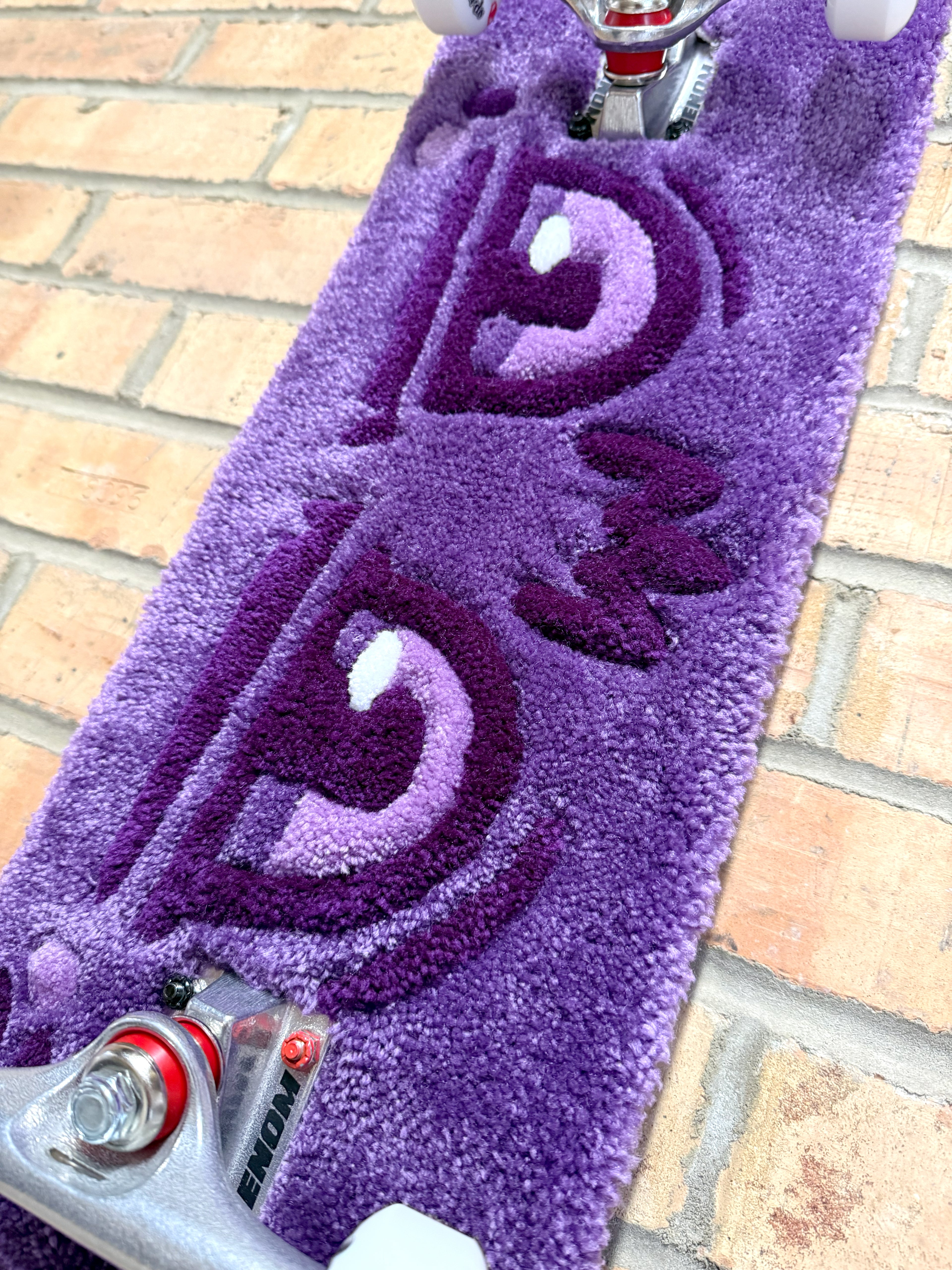 Close-up of the Dirty Look Rugboard featuring a handmade tufted purple eye design mounted on a skateboard deck with trucks and wheels.