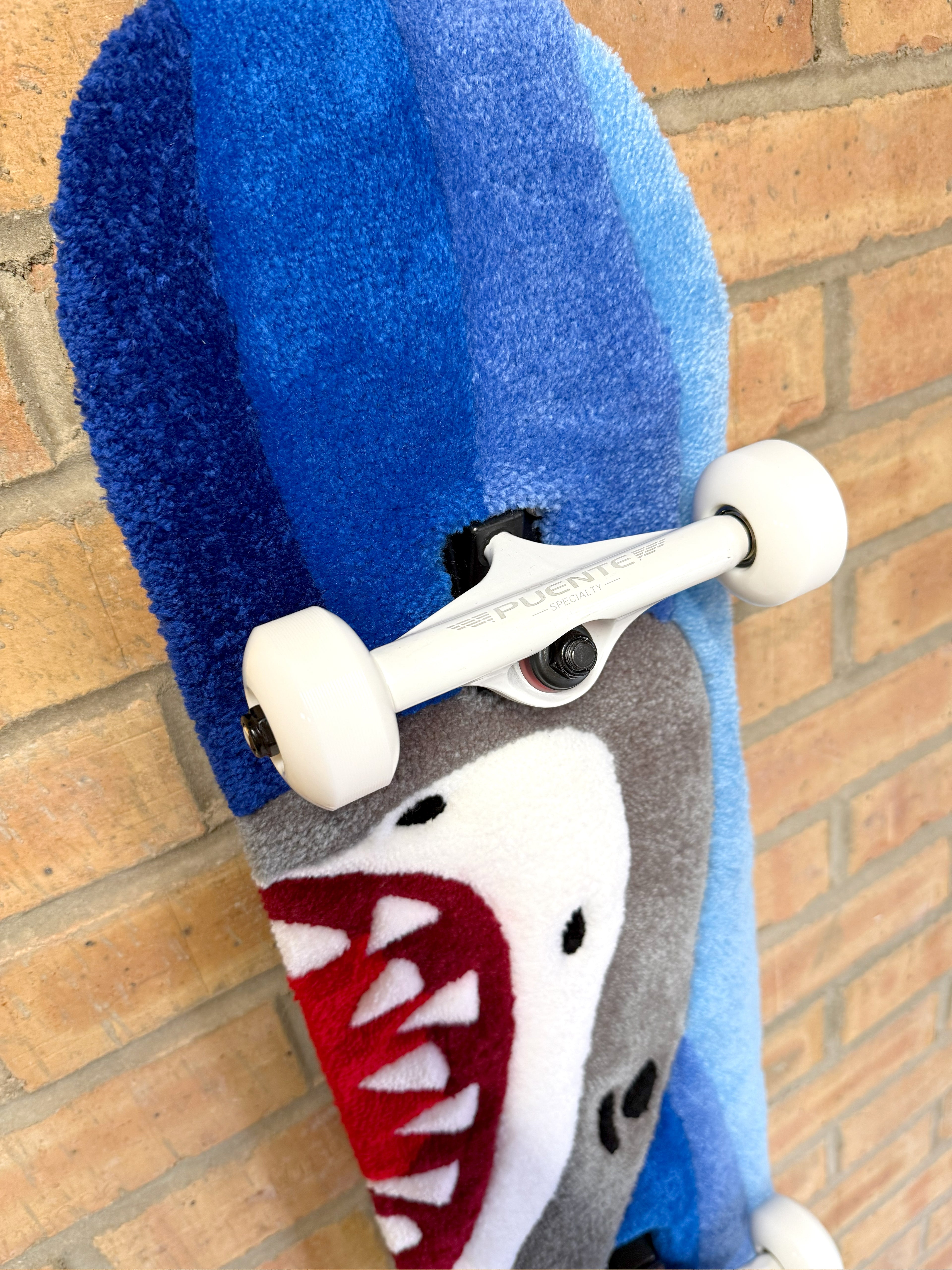 Close-up of the Shark Rugboard showing white skateboard hardware, blue ocean-inspired tufting, and shark graphic detail